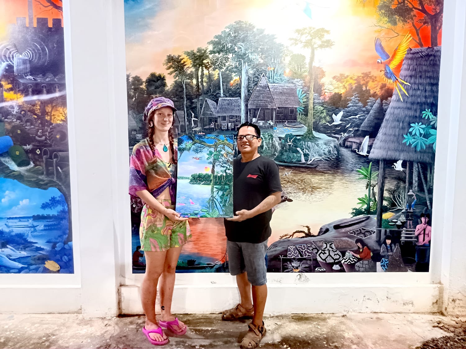 David Amaringo and a friend holding a painting in front of large wall murals depicting the Amazon river